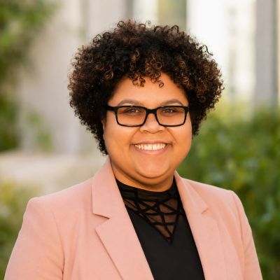 Smiling person with curly hair and glasses wearing a pink blazer.