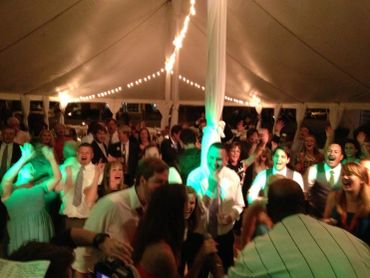 A lively crowd dancing and celebrating under a tent with string lights.