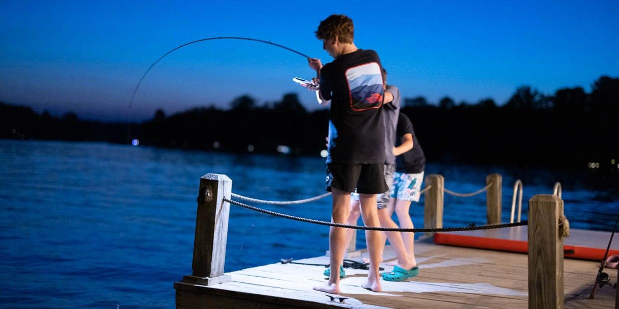 Three boys fishing on a dock by a lake at dusk.