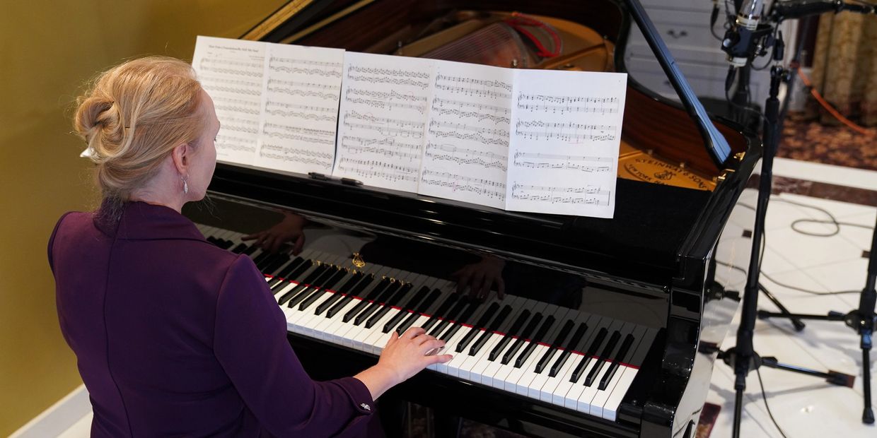 Woman playing a grand piano with sheet music and microphones set up nearby.