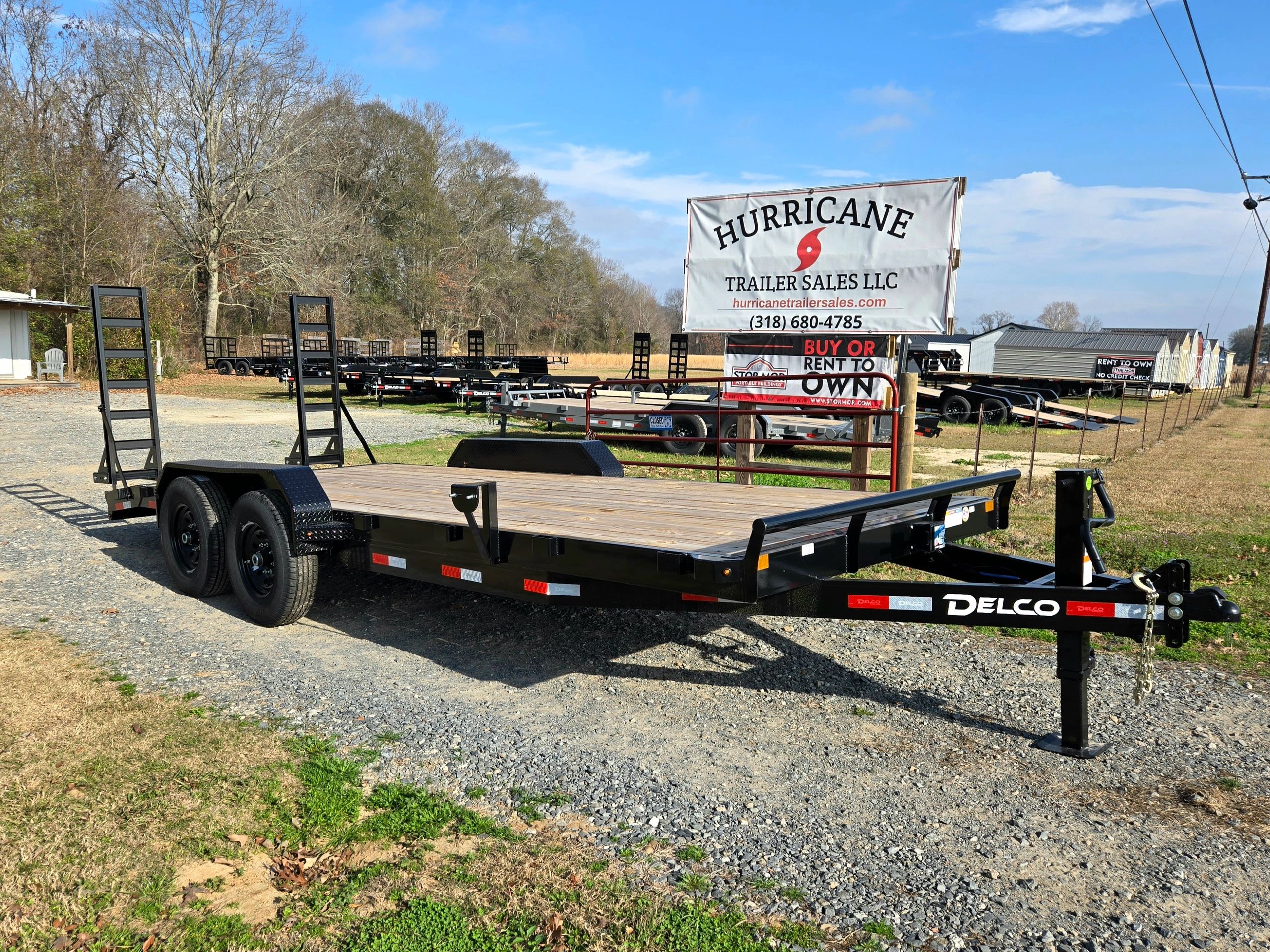 A flatbed trailer parked on a gravel lot with a sales sign in the background.