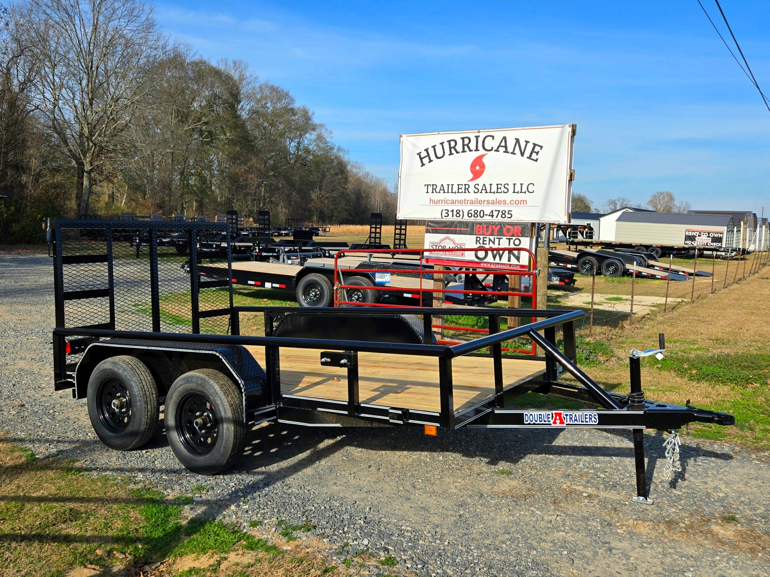 Black utility trailer with wooden bed at a trailer sales lot on a sunny day.