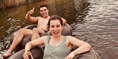 Two people relaxing on inner tubes in a calm river surrounded by tall grass.