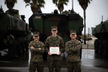 Three marines in uniform standing with a certificate, armored vehicles behind them.