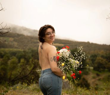Smiling woman with glasses holding a colorful bouquet in a natural outdoor setting.