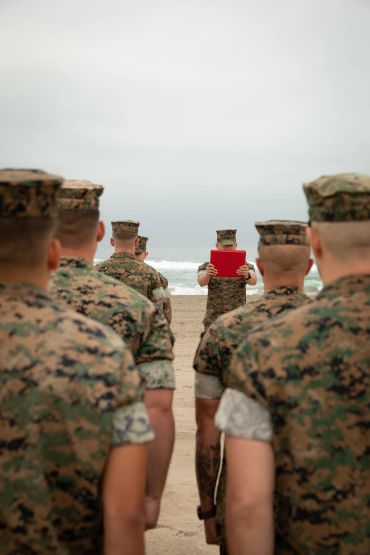 Marines in uniform standing on a beach, one holding a red folder.