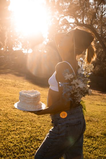 Pregnant woman holding a cake outdoors with sunlight behind her.