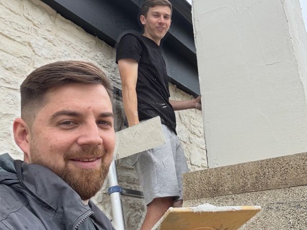 Two men plastering a wall, smiling at the camera.