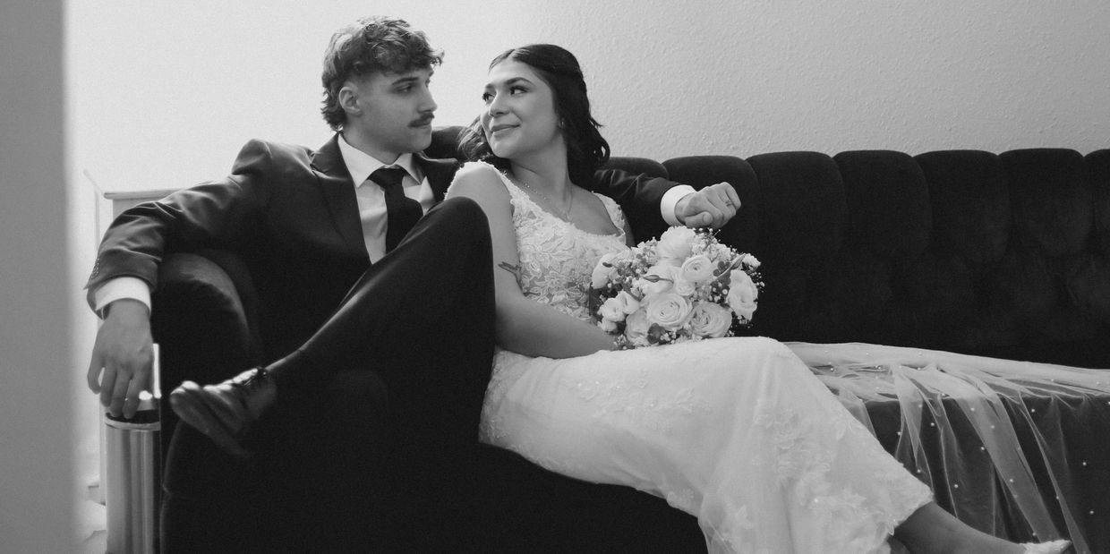 A newlywed couple shares a tender moment on a couch, the bride holding a bouquet.