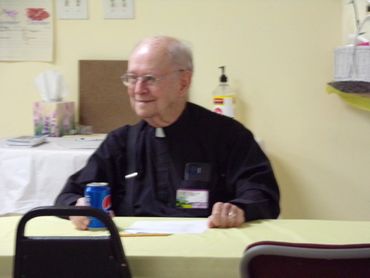 Elderly priest sitting at a table with a Pepsi can, smiling.