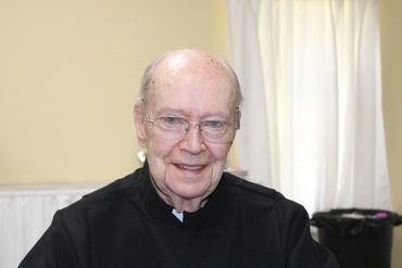 Elderly man smiling warmly in a black outfit indoors.