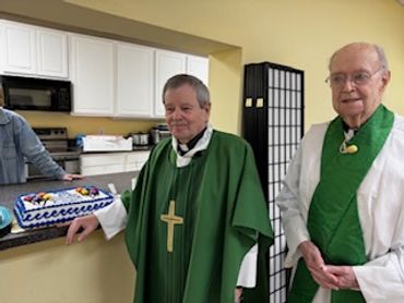 Two elderly men in green clerical robes standing near a decorated cake in a kitchen.