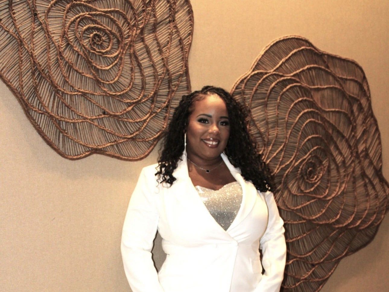 Smiling woman in white blazer with curly hair stands before decorative woven wall art.