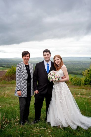 A bride, groom and Justice of the Peace standing together in front of a green meadow vista.