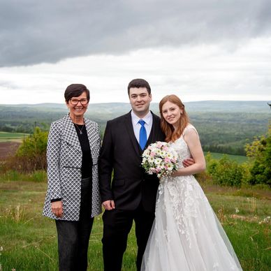 Bride, groom, Justice of Peace standing in front of panoramic field of green fields in Nova Scotia.