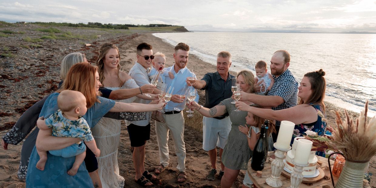 A family raising glasses of champagne to toast a newly married couple on a beach at sunset.