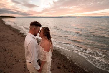 A bride and groom facing one another while standing on a beach in front of a pink sunset sky.