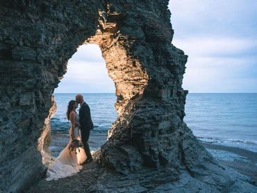 Nova Scotia elopement with bride and groom standing in a rocky arch at sunset on a beach.