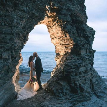 A bride and groom standing in a rocky arch with the ocean behind them.