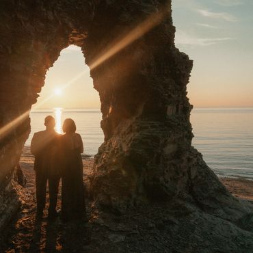 A bride and grooms silhouette against a golden sunset while standing under a rocky arch on a beach.
