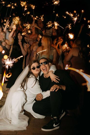 Bride and groom wearing sunglasses at night surrounded by friends waving sparklers all around them.