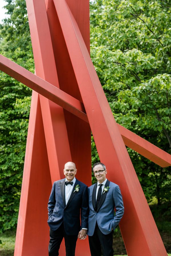 Two grooms on their wedding day standing in front of a red, modern, metal art structure in a forest.