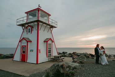 Nova Scotia elopement showing a couple standing beside a lighthouse and a pink sunset.