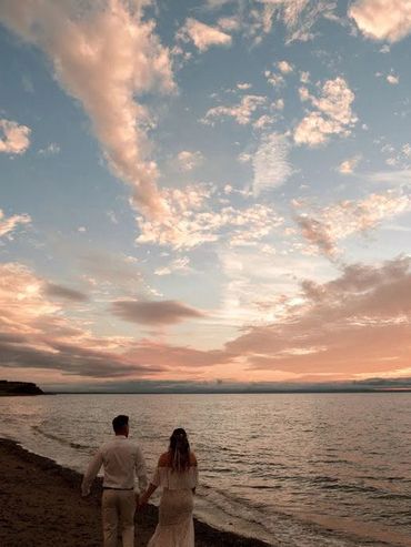 A bride and groom walking along the beach towards a purple pink sunset over the ocean.