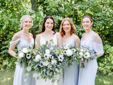 A bride and 3 bridesmaids standing together holding bouquets in front of a green forest backdrop.
