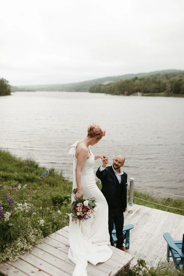 A bride and groom holding hands as they step onto a lakeside dock after their wedding.