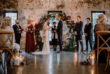 A bride, groom and Justice of the Peace stand with the wedding party in front of a flowery arch.