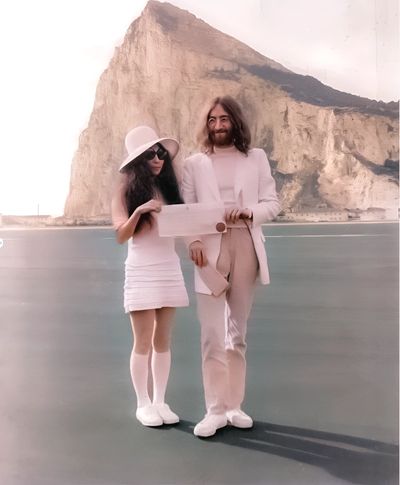 A stylish couple dressed in white stands near a large rocky cliff by the water.