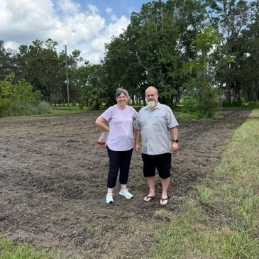 Older couple standing on freshly tilled soil with trees in the background.