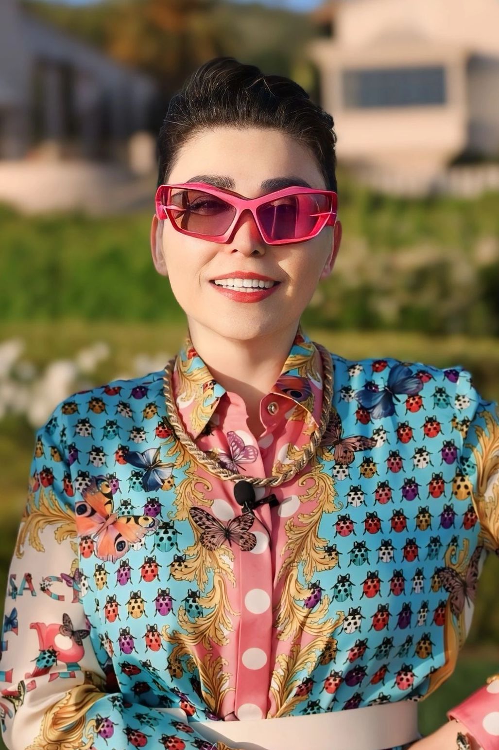 Woman smiling outdoors in colorful butterfly and ladybug patterned shirt with pink sunglasses.