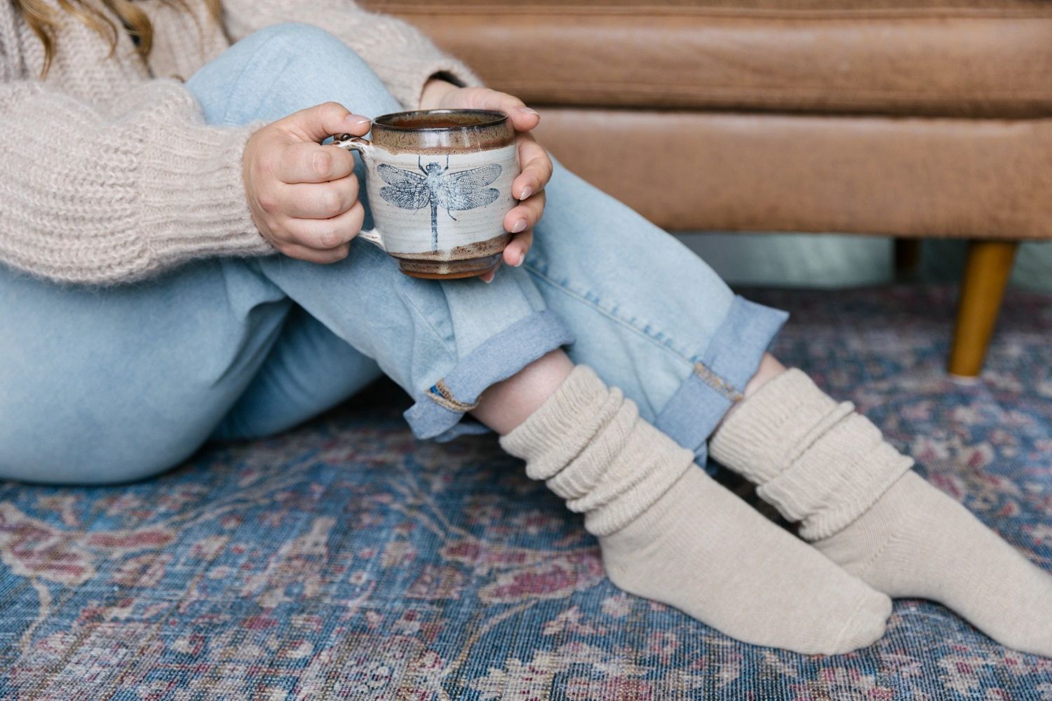 Person in cozy socks and sweater holding a mug with a dragonfly design.