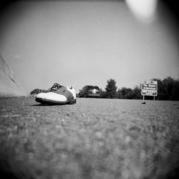 A lone shoe lies on a road with a car and trees in the background.