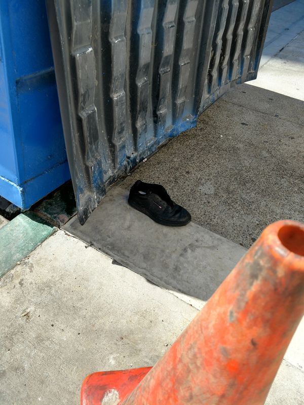 A single black shoe on a sidewalk near a blue dumpster and an orange traffic cone.