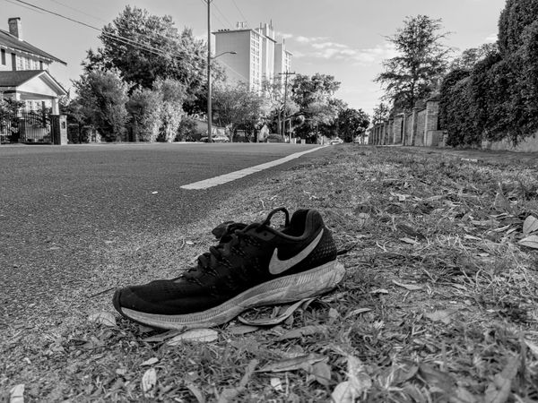 Black and white photo of a Nike shoe on the roadside with houses and trees in the background.