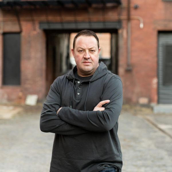Man in a black hoodie standing with arms crossed in an urban setting.