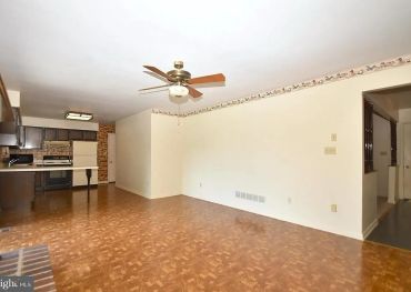 Empty living room with ceiling fan and kitchen in background.