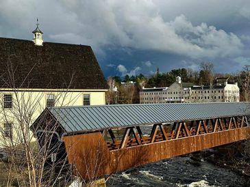 Wooden covered bridge over a river with buildings and cloudy sky.