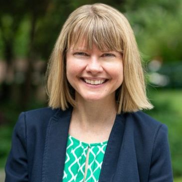 Woman in a blue jacket and green shirt smiling at the camera outside.