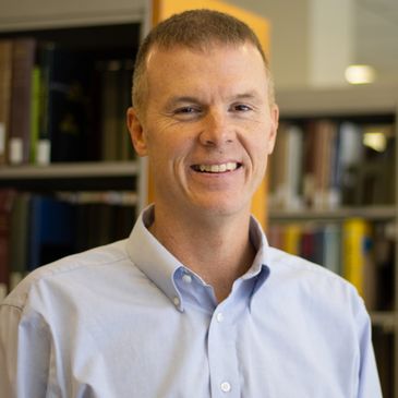Man standing in front of bookcases wearing a button down shirt and smiling at the camera.