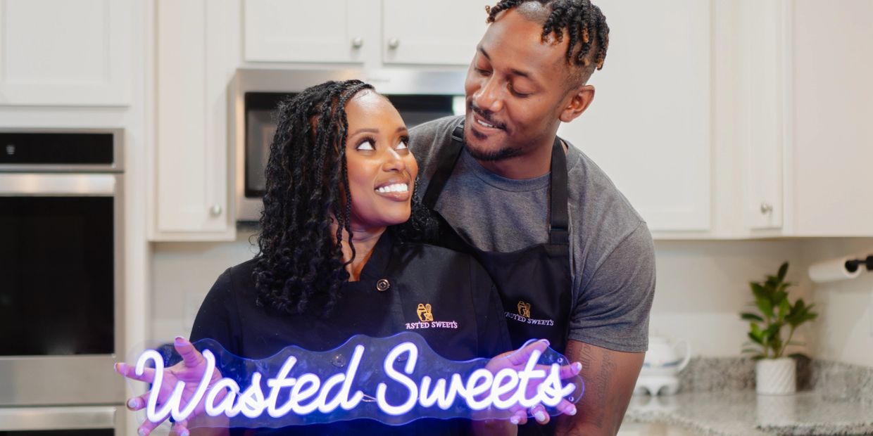 Couple holding a neon Wasted Sweets sign in kitchen.