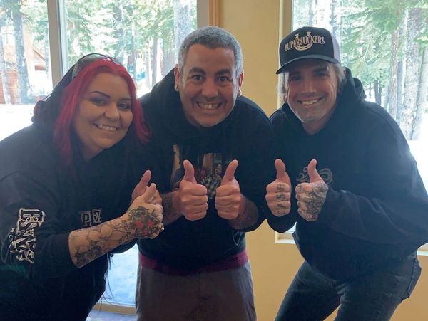 Three people smiling and giving thumbs up indoors with tattoos visible.