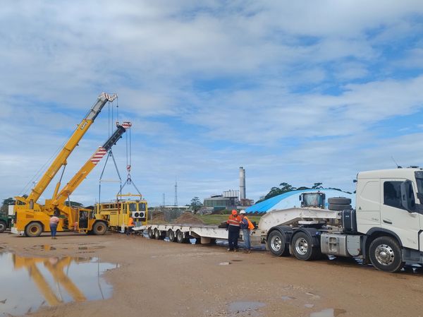 Two cranes lifting a yellow vehicle onto a flatbed truck at a construction site.