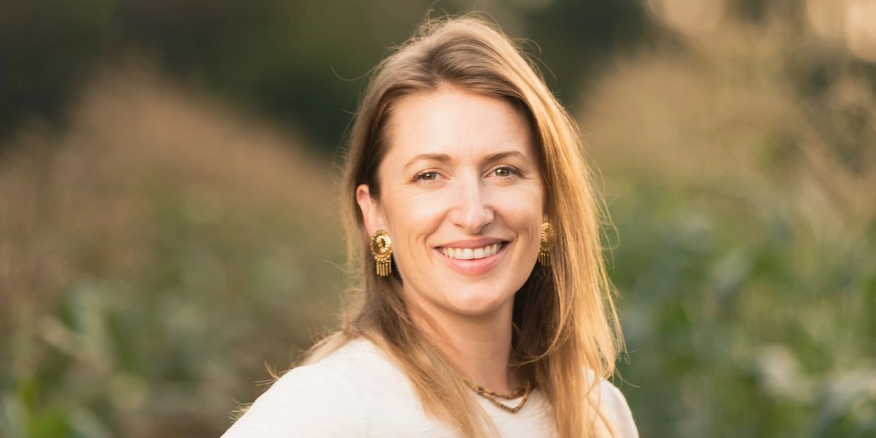 Smiling woman with blonde hair and gold earrings in a natural outdoor setting.