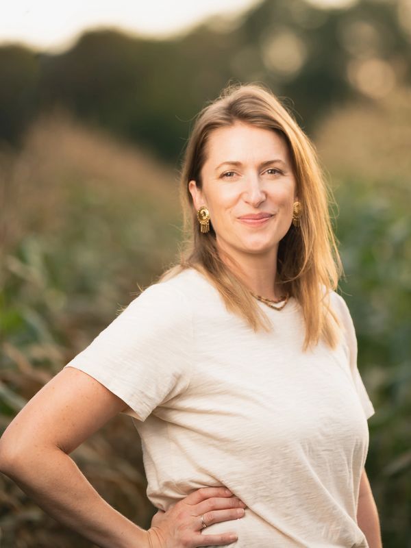 Confident woman smiling outdoors with hand on hip, wearing a beige shirt and gold earrings.