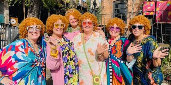 Six women in colorful clothes and orange wigs smiling and waving outdoors.