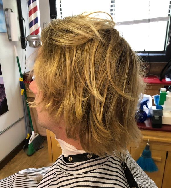 Side view of a person with messy blond hair in a barber's cape at a barbershop.